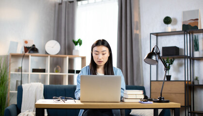 Attractive japanese female freelancer in casual outfit using modern laptop for work at home. Asian woman with dark hair sitting at table and typing on computer. Korean girl student studying at home