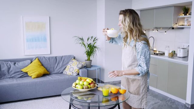 Pretty curly haired lady in grey apron pours fresh orange juice into cup from squeezer at glass table in light contemporary kitchen in morning