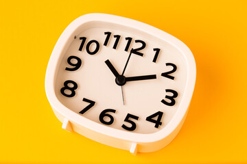 Small white alarm clock, black numbers, set the time placed on a table. Clock on isolated yellow background.