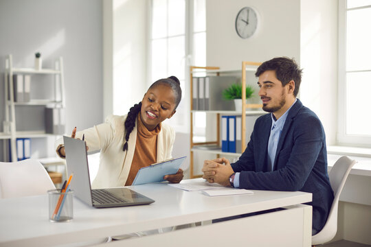 Happy Young Man Consulting Bank Manager Or Loan Advisor. Professional Real Estate Agent Meeting With Client, Giving Advice And Showing House Variants On Laptop Computer Screen Sitting At Office Desk