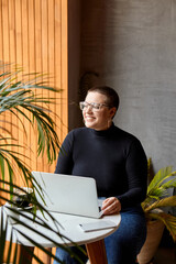 A young girl with glasses, short hair and a nose piercing is sitting in a cafe and working on a laptop. The concept of freelancing and remote work or training.