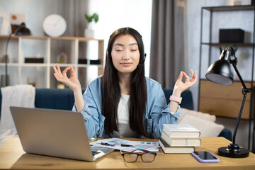 Asian woman freelancer in headset sitting at table with closed eyes and relieving stress by meditation at workplace. Concept of relaxation and harmony, no stress free relief at work