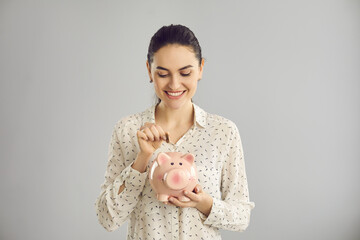 Happy beautiful independent young woman putting coin in piggy bank and smiling standing in studio...
