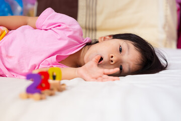 Obraz na płótnie Canvas Cute Asian girl lying around playing with toys on soft bed. Child made cheeky face and looked at camera. Kid leisure activities in the house. Lady wore a pink shirt. Children aged 4 years old.