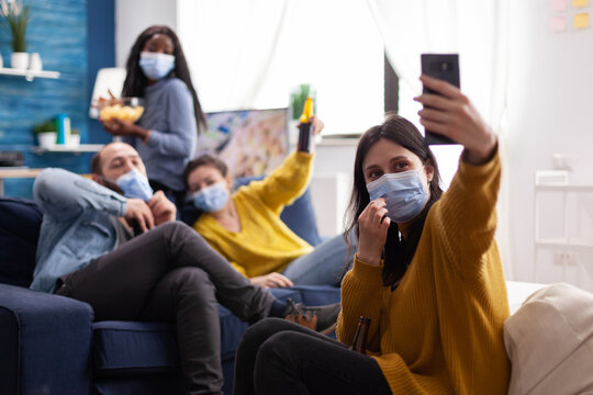 Cheerful happy mixed race friends taking selfie wearing face mask keeping social distancing wearing face mask against spread of virus sitting on couch in living room. Conceptual image.