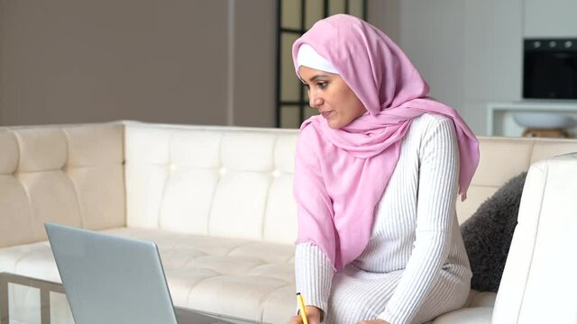 Female islamic student wearing hijab studying online, watching online webinar on the laptop and takes notes sitting on the couch at home, muslim woman remotely studying online. E-learning concept