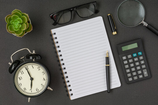Black vintage alarm clock, calculator, spectacles, notebook and a pen with a copy space on black background. Flat lay.