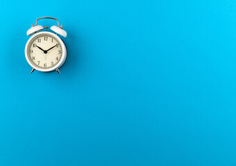 A white retro alarm clock shows ten hours and ten minutes on a blue paper background with a copy space. Flat lay. Top view. Place for text. The concept of summer, sunny and happy time. Time: 10.10