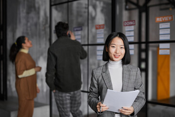 Waist up portrait of smiling Asian businesswoman standing by glass wall while planning project in modern office, copy space