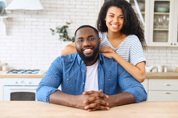 Portrait of African-American couple in love in the kitchen at home, a beautiful biracial woman hugs the shoulders of handsome black man, they look at the camera and smile spending time at home
