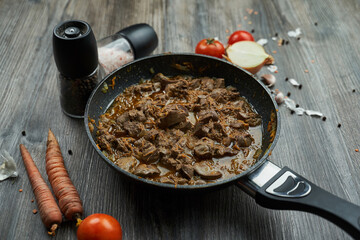 fried chicken liver with onions and carrots in a black pan on a wooden textured table. there are vegetables nearby