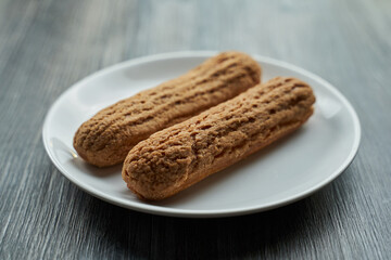 two eclairs with custard inside on a white plate. the plate stands on a gray wooden table.