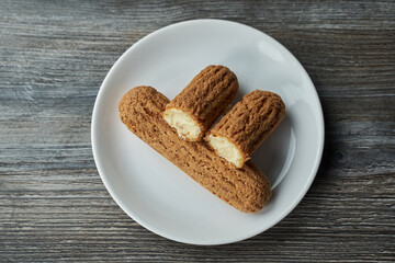 two eclairs with custard inside on a white plate. one is cut in half and lies on the second. the plate stands on a gray wooden table. view from above