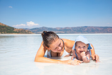 Young woman in swim suit and her daughter little baby girl are laying on white clay of turquoise crater lake Salda Golu, Turkey