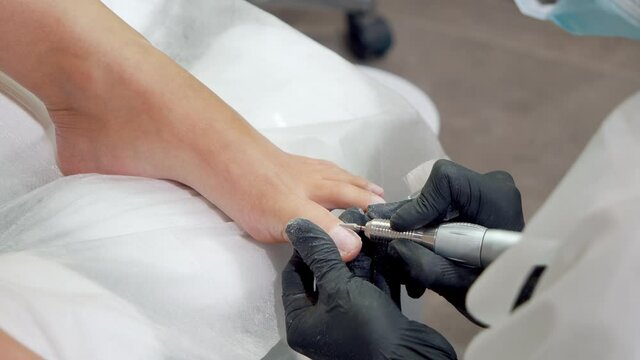 Close-up shot of hands in gloves doing a pedicure procedure with a special device in modern spa salon. Hardware medical pedicure with nail file drill apparatus.
