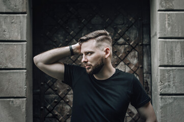 Male beauty, fashion. Portrait of a handsome hipster with beard and stylish haircut, dressed in a black  t-shirt, stands with a thinking look on a dark background.