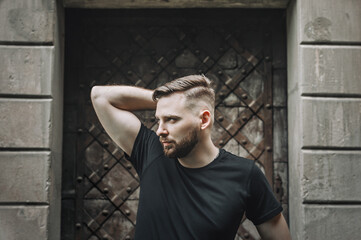 Side view portrait of thinking stylish young bearded man looking away. Portrait of a smart young man wearing a black t-shirt standing against against the background of old doors.