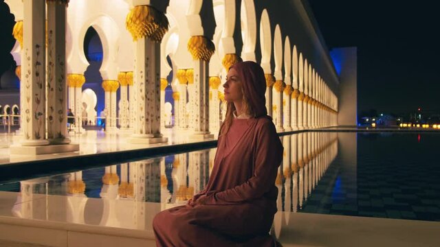 Portrait beautiful young woman tourist, pink muslim dress Abay. Girl sits on white marble bench. Beauty night lighting, columns reflected in water. UAE 4k 