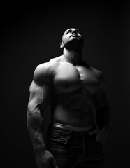 Fototapeta na wymiar Black and white. Strong muscular man, fitness trainer, bodybuilder stands in jeans and shirtless and looks up over dark background