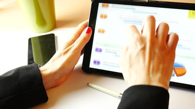 Business woman scrolling tablet with the hand, showing a gantt for project management purposes. Smartphone and pen on the desk