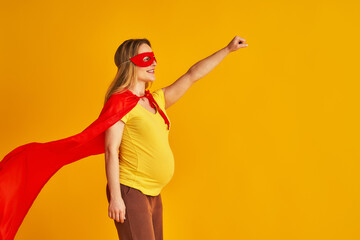 attractive smiling pregnant woman in superhero costume, wearing red mask and cape, stands with her arm outstretched on a yellow background. concept superpowers of a girl, feminism, desire to win