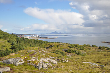 Fototapeta na wymiar View from Lovund hills to the rocky coastline of Lovundf and Lovund harbor on sunny summer morning. White clouds over the blue sea. Donna island on the horizon.