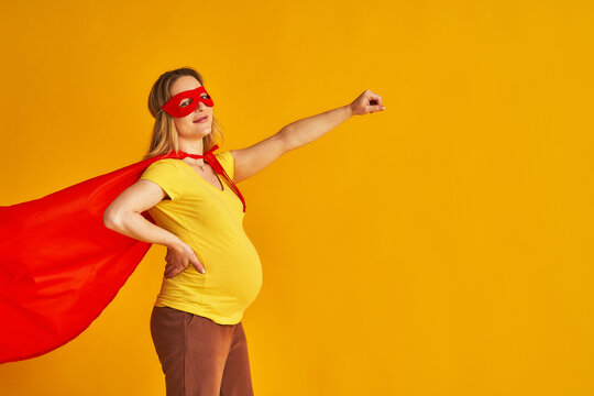 attractive smiling pregnant woman in superhero costume, wearing red mask and cape, stands with her arm outstretched on a yellow background. concept superpowers of a girl, feminism, desire to win
