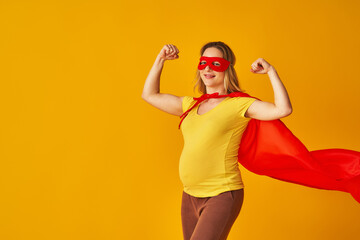 attractive smiling pregnant woman in superhero costume, wearing red mask and cape, showing her strength, muscles, stands on yellow background. concept superpowers girl, feminism, desire to win