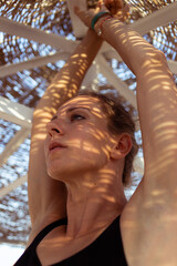 Fototapeta premium Beautiful woman in swimsuit standing under a beach umbrella with contrasting shadows from branches on the face. Summer vacation concept