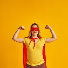 attractive smiling pregnant woman in superhero costume, wearing red mask and cape, showing her strength, muscles, stands on yellow background. concept superpowers girl, feminism, desire to win