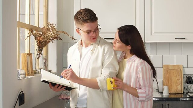 young man is standing in the kitchen making notes in a notebook, holding it in his hands, and his girlfriend is standing next to him and reading notes in a notebook.