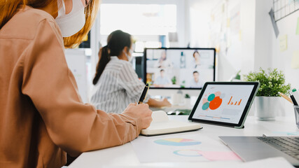 Asia businesspeople wear face mask using desktop talk to colleagues discussing brainstorm about plan in video call meeting in new normal office. Lifestyle social distancing and work after coronavirus.