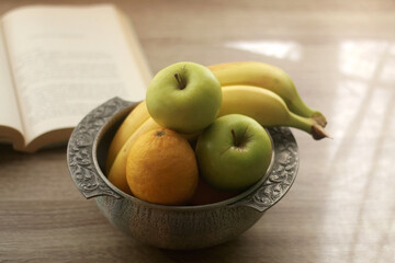 Antique bowl with bananas, apples and lemons and open book on wooden table. Selective focus.