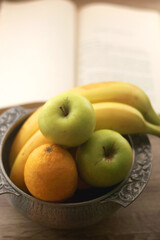 Antique bowl with bananas, apples and lemons and open book on wooden table. Selective focus.