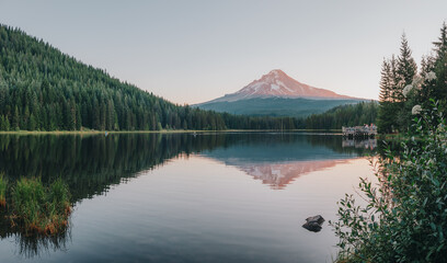 Panoramic landscape of the sunrise glow on the peak of Mount Hood and the perfect glass reflections, summer wildflowers and the dock over Lake Trillium in Mount Hood National Forest, Oregon, USA. 