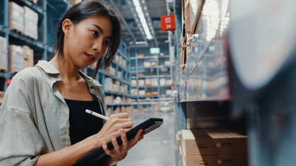 Attractive young Asia businesswoman manager looking for goods using digital tablet checking inventory levels standing in retail shopping center. Distribution, Logistics, Packages ready for shipment.