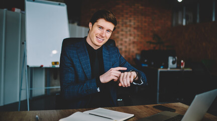 Portrait of cheerful male employer in elegant formal wear smiling at camera while checking clock time, successful Caucasian businessman using modern smartphone during work day in office interior