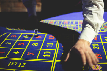 A close-up vibrant image of multicolored casino table with roulette in motion, with casino chips. the hand of croupier, money and a group of gambling rich wealthy people playing in the background