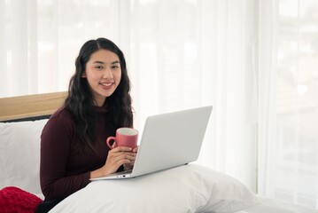 Asian woman in casual outfit sitting on bed using laptop and drink hot beverage in white modern bedroom