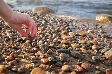 Hand takes one small stone among the many lying on the sandy bank of the river, against the background of water on a spring.
