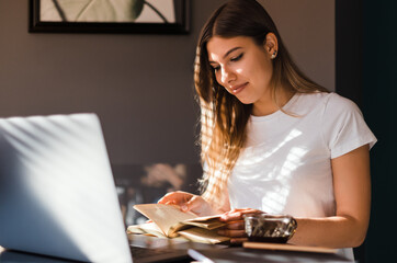 Happy young caucasian woman reading book in front laptop at the kitchen in sunny morning and drinking coffee.