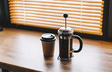 Coffee brewed in a french press and paper cup on a wooden table near window.
