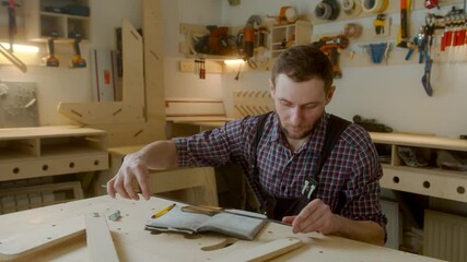 curly joiner master using smartphone checks measurements , handsome Caucasian young man carpenter in apron and caged shirt holding mobile device sitting at work-desk - Powered by Adobe