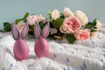Two pink Easter eggs with bunny ears close-up on a white knitted blanket and a blue background with flowers