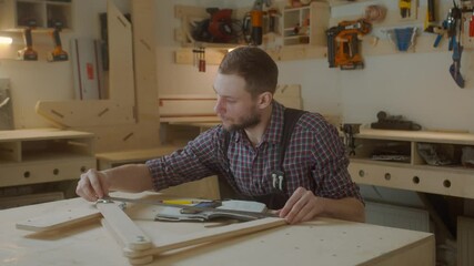 curly joiner master using smartphone checks measurements , handsome Caucasian young man carpenter in apron and caged shirt holding mobile device sitting at work-desk - Powered by Adobe