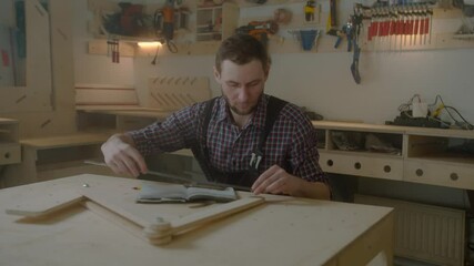 curly joiner master using smartphone checks measurements , handsome Caucasian young man carpenter in apron and caged shirt holding mobile device sitting at work-desk - Powered by Adobe