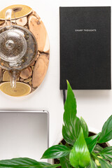 glass teapot with herbal tea on wooden stand, gray tablet, green plant and notebook on white wooden table