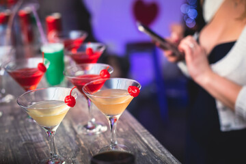 Female lonely girl glam blond woman using smartphone with dating app site is drinking in a bar on a counter with different coloured alcohol cocktail drinks setting on bar counter in the night club