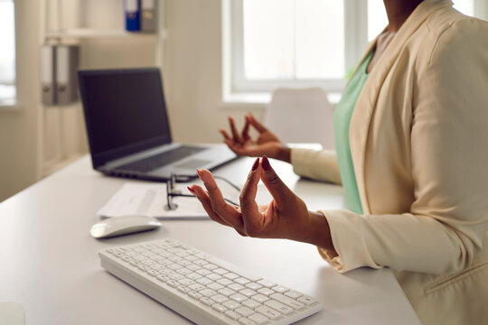 Office worker relieving stress during work day. Calm young black woman sitting at desk with laptop computer, doing Gyan Mudra, meditating, relaxing and reaching zen, closeup. Body and mind health care