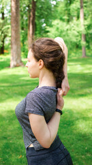 Yoga stretch exercise. Young brunette woman doing back hands lock for spine health in park, standing on green grass background. Training class outdoor. Workout for beautiful posture. Vertical